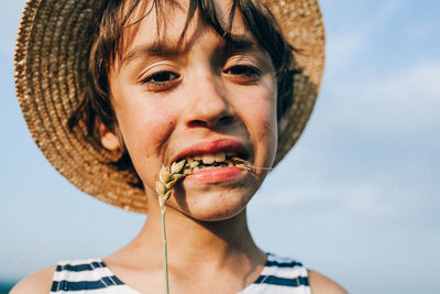 Portrait of boy wearing hat biting crop against sky