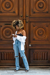 Full body of african american female in stylish casual outfit standing near entrance and surfing cellphone