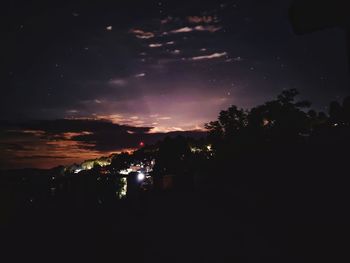 Silhouette trees and illuminated buildings against sky at night