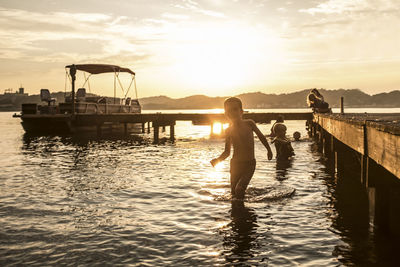 A boy walking out of the water at a lake with sunset and boat behind