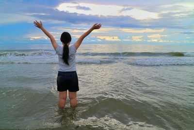 Rear view of woman with arms raised standing in sea during sunset