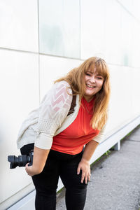 Portrait of smiling young woman holding camera standing against wall outdoors