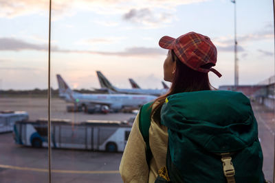 Back view of young asian female traveler with backpack standing near window in airport terminal and observing airplanes while waiting for flight in sri lanka
