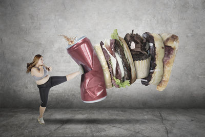 Overweight woman kicking drink can and fast food against wall