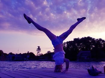 Full length of young woman doing headstand on pier over lake against cloudy sky during sunset