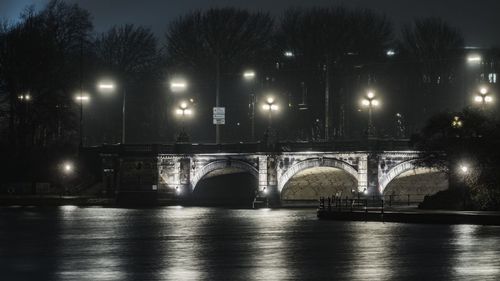 Illuminated bridge over river against trees at night