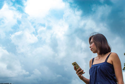 Low angle view of thoughtful woman using phone while standing against cloudy sky