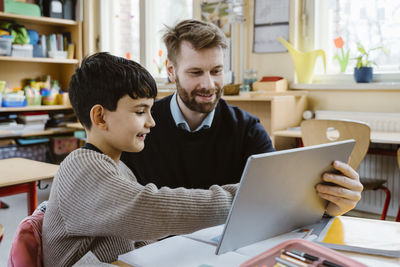 Smiling male teacher assisting schoolboy using tablet pc at desk in school