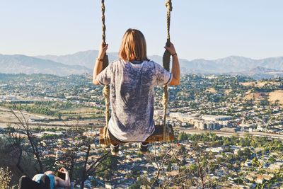 Rear view of woman sitting on swing against cityscape