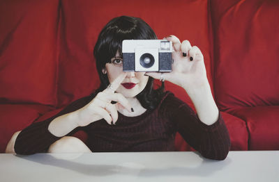 Woman covering face with analog camera in front of red couch