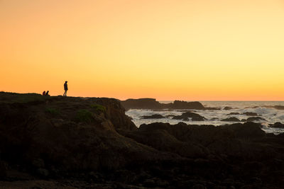 Silhouette people on rocky shore against clear orange sky