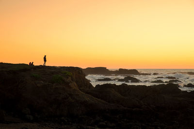Silhouette man standing on cliff by sea against orange sky