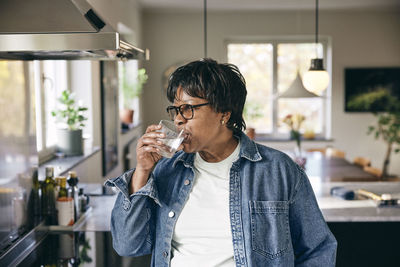 Senior woman wearing denim jacket drinking water in kitchen at home