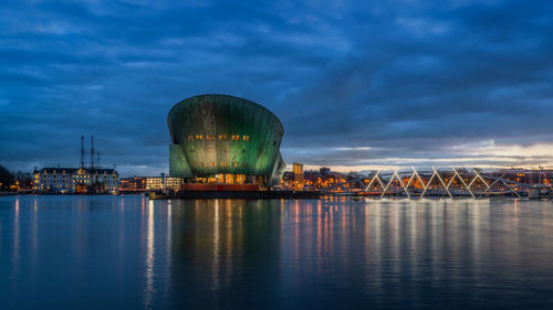 Illuminated city by sea against cloudy sky