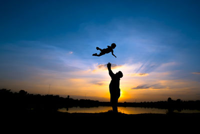 Silhouette father throwing son into air while standing at lakeshore against sky during sunset