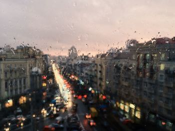 Illuminated cars on road amidst buildings seen from wet glass window