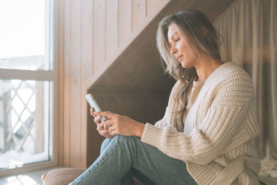 Young woman with blonde long hair in cozy knitted white sweater with mobile phone in hands at home