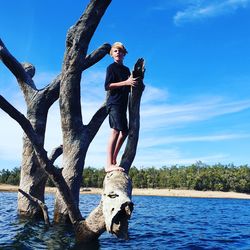 Teenage boy standing on bare tree over lake against blue sky