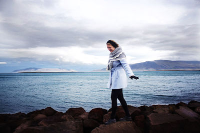 Woman walking on rocky shore against cloudy sky