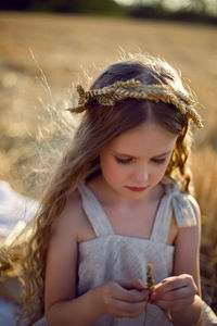 Girl child in a dress and a wreath on her head sit on a mown field of wheat at sunset in summer