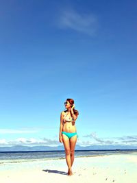 Full length of woman in bikini standing at beach against blue sky