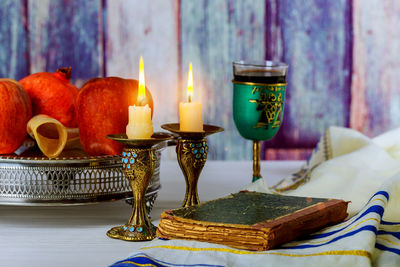 Burning candlestick holder by book and fruits on table