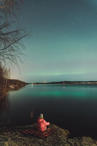 Back view of unrecognizable explorer sitting on shore of lake at night and enjoying spectacular scenery of northern lights in winter