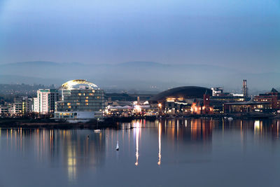 Illuminated cityscape by sea against sky during sunset