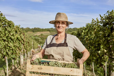 Smiling mature farmer wearing straw hat holding crate of fresh grapes in vineyard