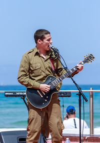 Young man playing guitar at sea against clear sky