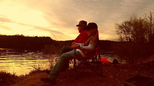 Men sitting by lake against sky during sunset