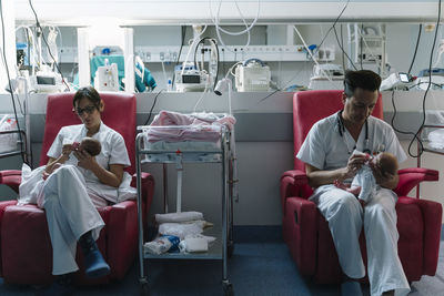 Doctors sitting on armchairs and feeding newborns from bottles holding heads in room with equipment in hospital