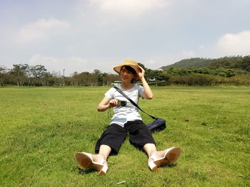 Portrait of woman photographing while sitting on field against sky