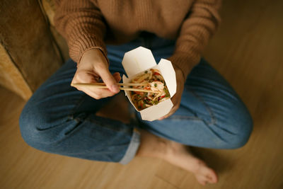 Girl in casual clothes eating chinese noodles from a box sitting on the floor, food delivery