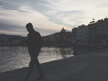 Silhouette man standing by river in city against sky during sunset