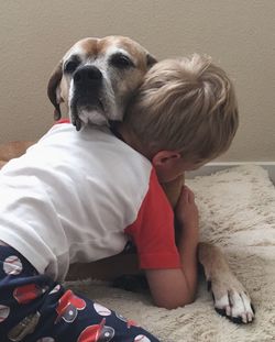 Rear view of boy embracing dog on rug at home