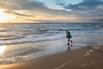 Boy running on shore at beach against sky during sunset