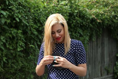 Young woman closing lipstick while standing against ivy wall during sunny day