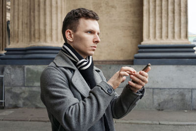 Portrait caucasian young man using his mobile phone in gray coat stands