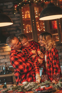 Smile active parents with small son in red checkered sleepwears waiting santa indoor.