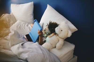 High angle view of boy lying with stuffed toy while using digital tablet on bed at home