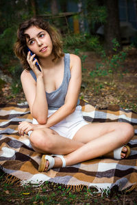 Beautiful teenager girl talking by phone and at the forest. young girl sitting on blanket at grass