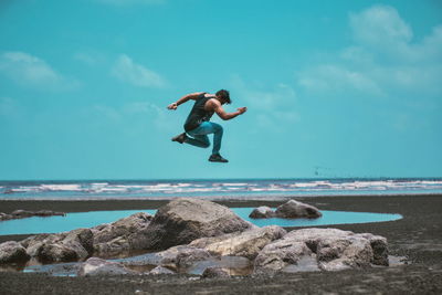Man jumping on rock at beach against sky