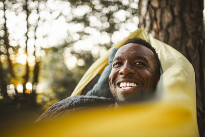 Smiling man looking away while lying over hammock in forest