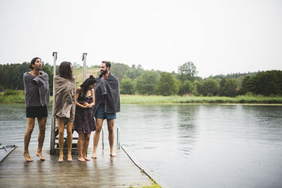 Friends wrapped in towels talking while standing on jetty over lake against clear sky