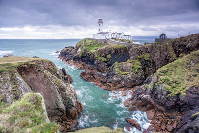 Lighthouse on cliff by sea against cloudy sky