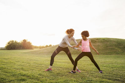 Mother assisting daughter in exercise on grass at park against sky