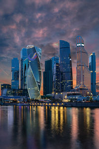 Illuminated modern buildings by bay against sky