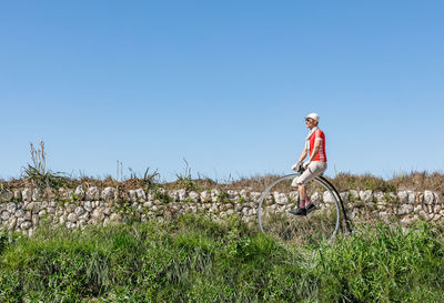 Side view of adult male in sunglasses and stylish casual clothes riding retro high wheel bike along footpath against green grassy field in summer day