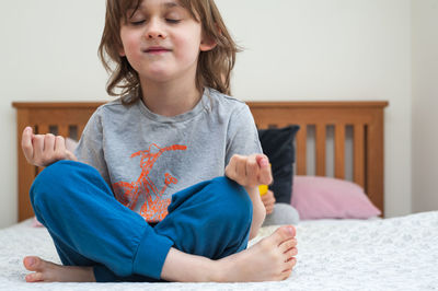 Cute, little boy sitting comfortably on bed in bedroom and training yoga in lotus pose.